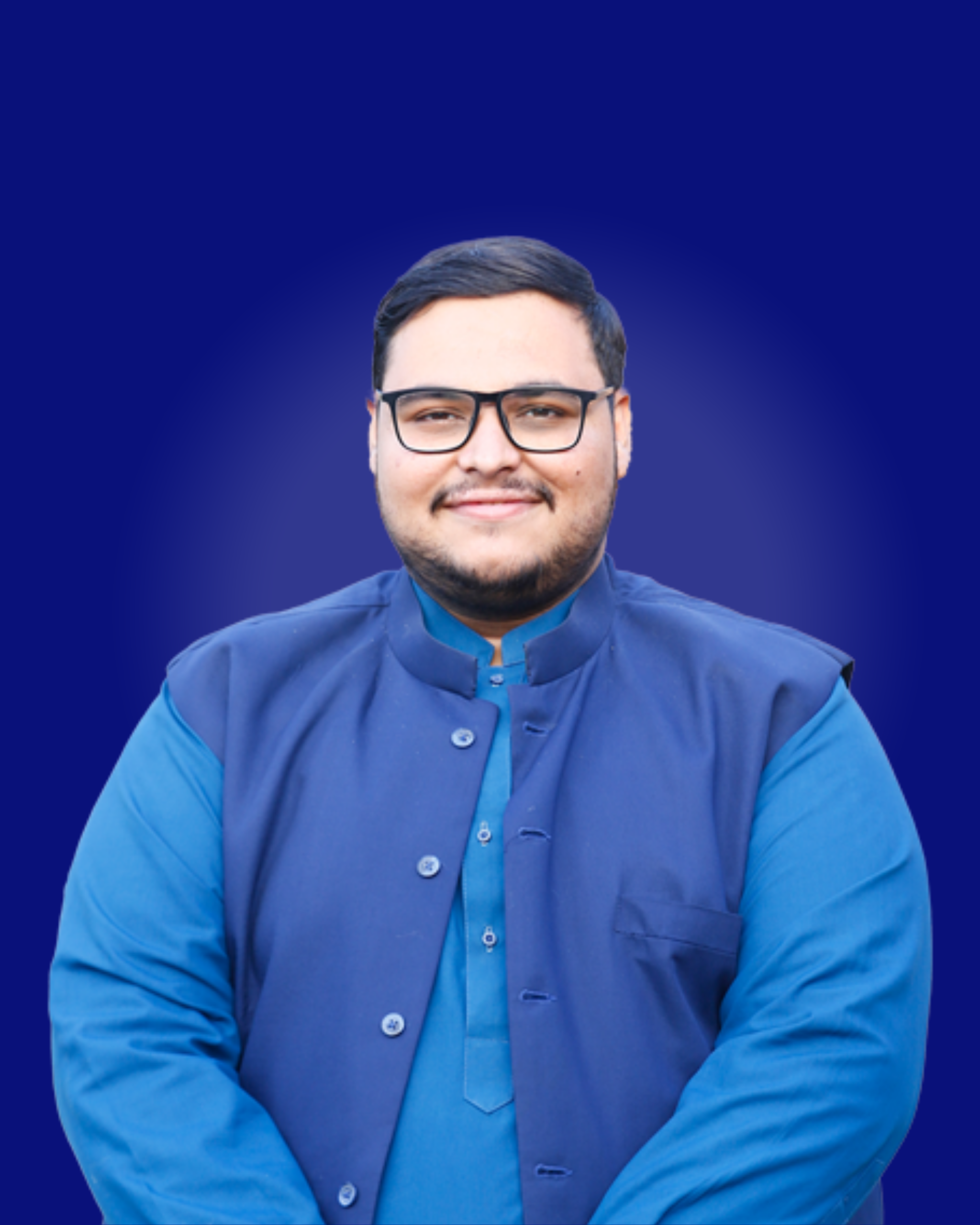 About Us A portrait of a young man with a beard and glasses, smiling confidently. He is wearing a traditional blue shalwar kameez topped with a dark blue vest (waistcoat/sadri). The background is a solid, deep blue with a light radial gradient centered on his face.