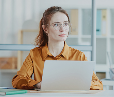 "Digital Marketing Agency"A headshot of a professional woman with her hair pulled back, wearing a mustard-yellow blouse and round glasses, seated at a desk. She is looking thoughtfully off-camera, with a silver laptop open in front of her. The background is a bright, modern office with glass partitions.