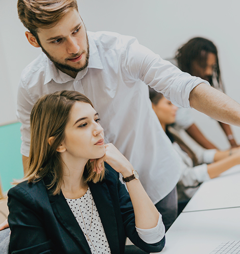 A focused shot of a diverse group of professionals working in an office. A young man in a white shirt is standing over a woman seated at a desk, pointing and discussing something on a screen, suggesting mentoring or a work review. Other colleagues are visible but out of focus in the background.